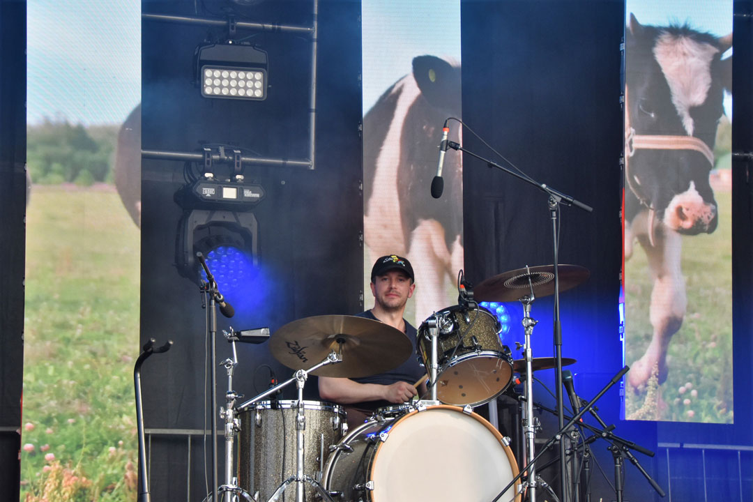 A drummer plays his drum set on stage with three tall video wall panels in the back ground