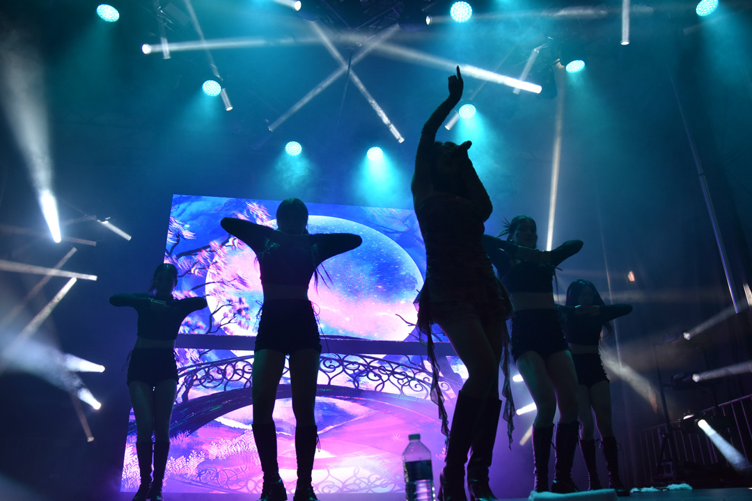 The silhouettes of five women performing are lit up by blue lights and a large LED video wall showing a pink and purple moon.