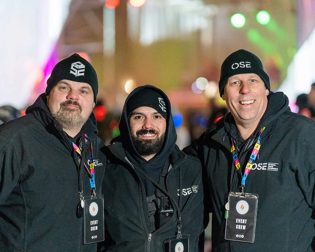 Three OSE crew members wearing branded jackets and winter hats smile for the camera during the Fire & Ice festival
