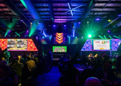 OSE Event Portfolio Photos - CANSEC - Reception - Set Design with Video Wall & Stage Lighting Design. Angled video walls displays on either side of the stage. Blue, green and purple lights lighting the stage and ceiling.